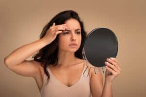 Concerned young woman looking in mirror and touching her forehead, illustrating early signs of aging and potential use of Jeuveau for facial rejuvenation